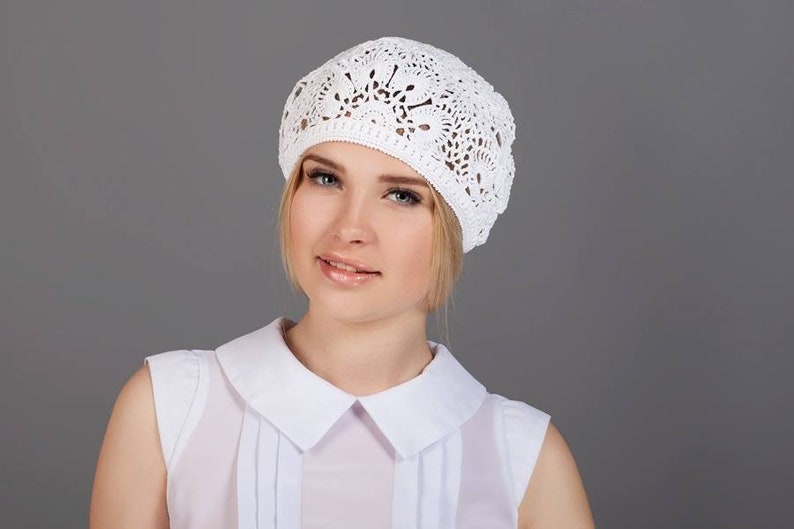 May include: A white crocheted beret with a delicate floral pattern. The beret is worn by a person with blonde hair and a white blouse.