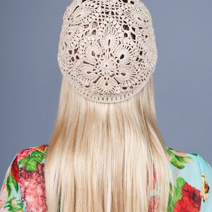 May include: A beige crocheted hat with an openwork design. The hat is worn on a person with long blonde hair. The person is wearing a floral patterned top with red and pink flowers.