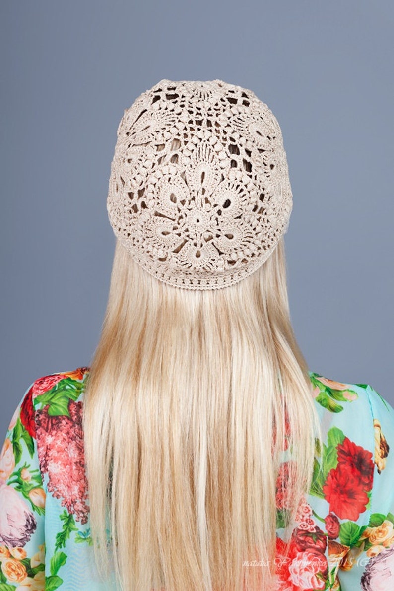 May include: A beige crocheted hat with an openwork design. The hat is worn on a person with long blonde hair. The person is wearing a floral patterned top with red and pink flowers.