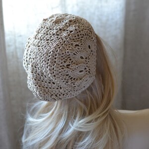 May include: A beige crocheted beret with a delicate openwork pattern. The beret is worn on a mannequin with long blonde hair.
