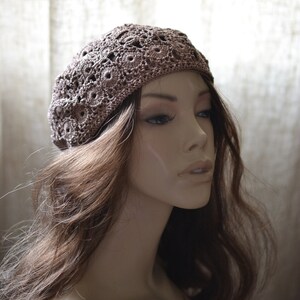 May include: A brown crocheted beret with a delicate floral pattern. The beret is worn on a mannequin with long brown hair.