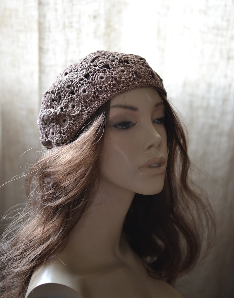 May include: A brown crocheted beret with a delicate floral pattern. The beret is worn on a mannequin with long brown hair.