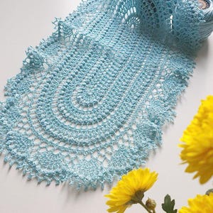 Puede incluir: Un tapete de ganchillo azul claro hecho a mano con un diseño calado delicado. El tapete está parcialmente enrollado, revelando una madeja de hilo a juego. Los crisantemos amarillos están en primer plano. El intrincado patrón del tapete es visible.