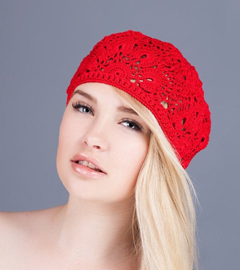 May include: A woman with blonde hair is wearing a red crocheted beret. The beret has a delicate floral pattern.