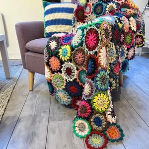Könnte beinhalten: Eine farbenfrohe, handgefertigte Häkeldecke mit floralem Muster, die über einem lila Sessel drapiert ist. Die Decke zeigt verschiedene Farben, darunter Rosa, Gelb, Blau und Grün. Ein blau-weiß gestreiftes Kissen liegt auf dem Stuhl.