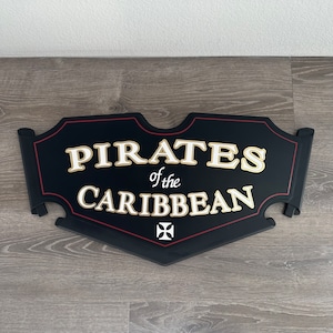May include: Black wooden sign with red trim and gold lettering that reads "Pirates of the Caribbean". A white cross is in the center of the sign.
