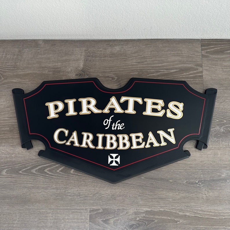 May include: Black wooden sign with red trim and gold lettering that reads "Pirates of the Caribbean". A white cross is in the center of the sign.