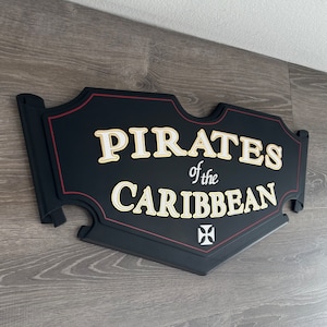 May include: Black wooden sign with red trim and gold lettering that reads "Pirates of the Caribbean". A white cross is in the bottom right corner.
