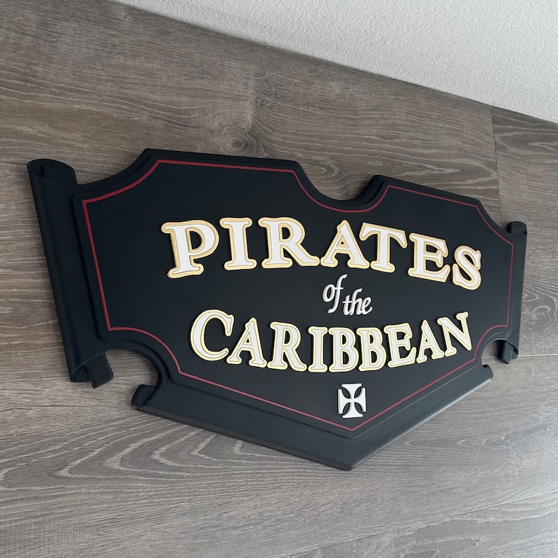 May include: Black wooden sign with red trim and gold lettering that reads "Pirates of the Caribbean". A white cross is in the bottom right corner.