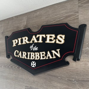 May include: Black wooden sign with red trim and gold lettering that reads "Pirates of the Caribbean". A white cross is in the lower center of the sign.