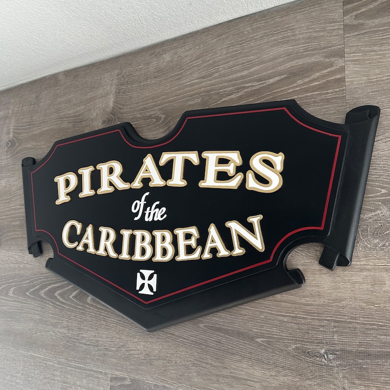 May include: Black wooden sign with red trim and gold lettering that reads "Pirates of the Caribbean". A white cross is in the lower center of the sign.
