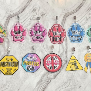 May include: Nine colourful paw print shaped badges with different messages about pronouns, photos, and dog behaviour. The badges are attached to silver clips. The messages include "Any Pronouns", "She/Her", "She/They", "He/Him", "He/They", "They/Them", "Overstimulated Proceed with Caution", "Ask for Photos", "Paws Off", "Warning Furry Trash", and "Furry Handler".