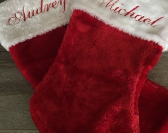 Puede incluir: Dos calcetines navideños rojos con puños de piel sintética blanca. El nombre "Audrey" está bordado en rojo en un calcetín y "Michael" en el otro. Los calcetines están hechos de un material suave y afelpado.