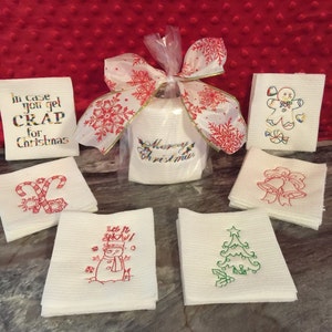 May include: A collection of white Christmas-themed embroidered washcloths. Designs include a gingerbread man, candy canes, a Christmas tree, bells, and a snowman. A roll of toilet paper is wrapped with a snowflake ribbon and the words "Merry Christmas".