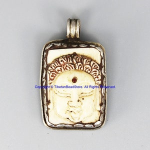 May include: A silver pendant with a carved bone Buddha face. The Buddha has a red dot on its forehead and is surrounded by a silver frame.