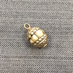 May include: A gold-colored, egg-shaped pendant with intricate filigree and a small loop at the top. The pendant has a cream-colored center and ornate detailing, suggesting a vintage or antique style. The background is a textured gray fabric.