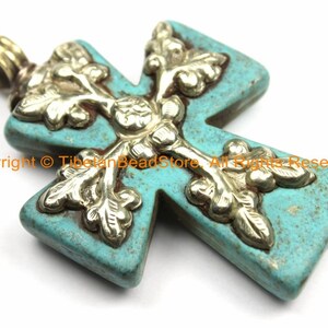 May include: A turquoise cross pendant with ornate silver-colored floral detailing. The pendant has a bail for hanging and is set against a white background. The text "Copyright © TibetanBeadStore. All Rights Reserved." is visible.