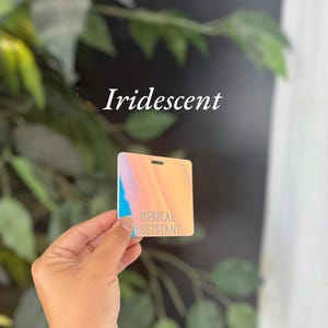 May include: A square, iridescent badge with a smooth finish, featuring the text "MEDICAL ASSISTANT" printed in a clear, sans-serif font. The badge displays a gradient of soft orange, pink, and blue hues, with a small rectangular slot at the top.