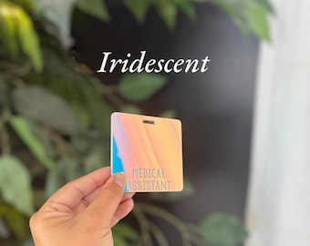 May include: A square, iridescent badge with a smooth finish, featuring the text "MEDICAL ASSISTANT" printed in a clear, sans-serif font. The badge displays a gradient of soft orange, pink, and blue hues, with a small rectangular slot at the top.