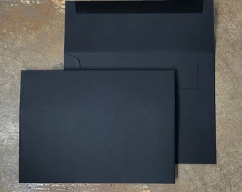 May include: Two black envelopes, one slightly larger than the other, are stacked on a textured surface. The larger envelope has a black flap and a black strip across the top.