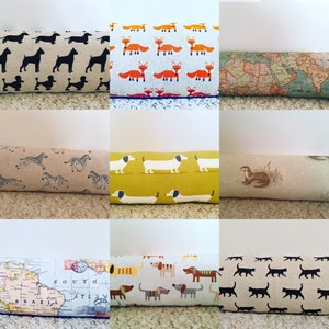 May include: A collection of decorative door draft stoppers. The stoppers feature various patterns, including black dog silhouettes, orange foxes, a world map, zebras, dachshunds, an otter, and black cat silhouettes. Each stopper is rectangular.