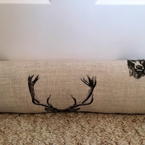 May include: A beige fabric door draft stopper with black deer antler and head illustrations. The rectangular pillow is designed to block drafts and features a rustic, country-inspired design.