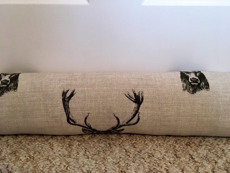May include: A beige fabric door draft stopper with black deer antler and head illustrations. The rectangular pillow is designed to block drafts and features a rustic, country-inspired design.