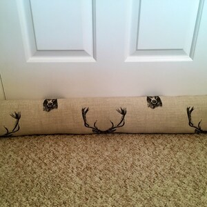 May include: A beige fabric door draft stopper with black deer antler and bear head designs. The long, cylindrical pillow is placed at the base of a white door on a beige carpet.