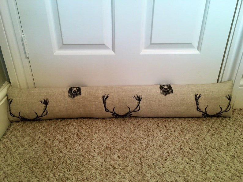 May include: A beige fabric door draft stopper with black deer antler and bear head designs. The long, cylindrical pillow is placed at the base of a white door on a beige carpet.