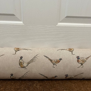 May include: A fabric door draught excluder in beige, patterned with pheasants in brown, tan, and black. The cylindrical stopper is placed against a white door. The pheasants are in various poses.