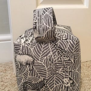 May include: A fabric doorstop in grey and white, decorated with a leaf design and safari animal illustrations. The animals include a giraffe, zebra, and lion. It has a handle and a tag that reads "Handmade by Hannah Jacobs Designs."