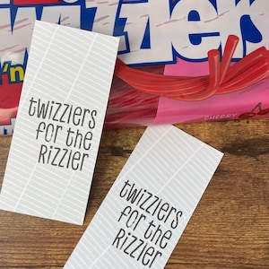 May include: Two white gift tags with black text that reads "Twizzlers for the Rizzie" on a wood background. The tags are printed with a white and gray striped pattern.