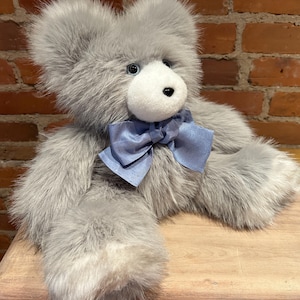 K&ouml;nnte beinhalten: Ein grauer Pl&uuml;schteddyb&auml;r mit einer hellblauen Schleife aus Satin. Der B&auml;r sitzt auf einer Holzoberfl&auml;che.