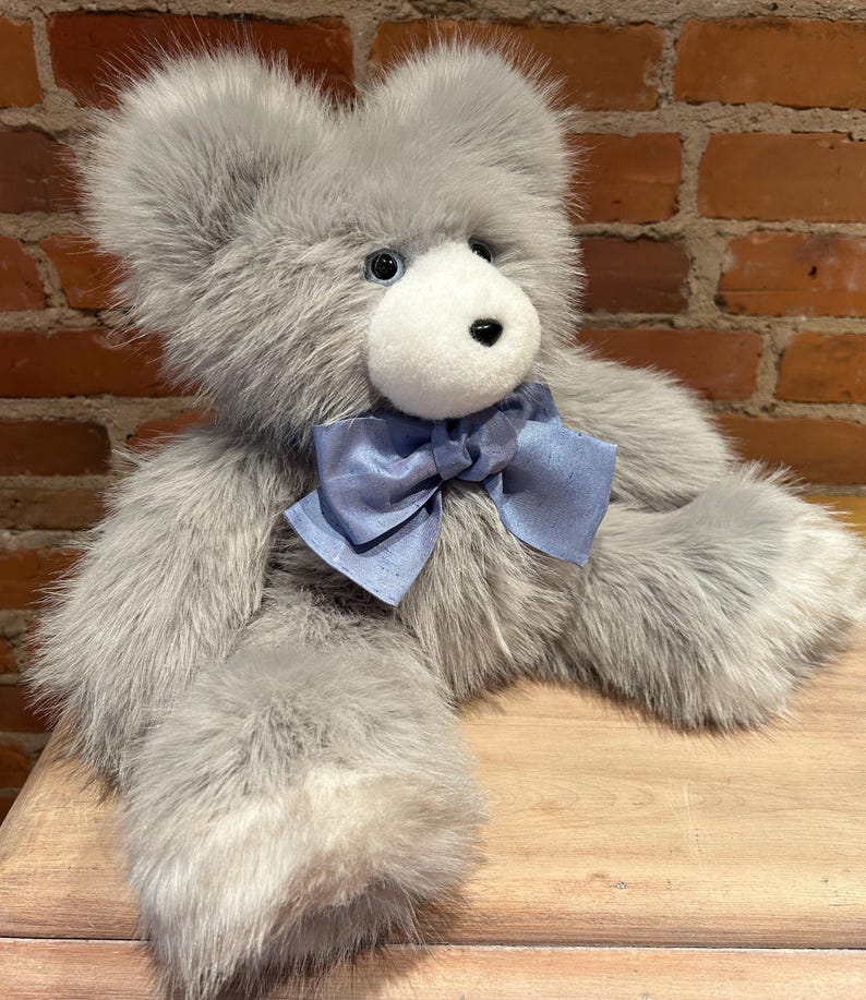 K&ouml;nnte beinhalten: Ein grauer Pl&uuml;schteddyb&auml;r mit einer hellblauen Schleife aus Satin. Der B&auml;r sitzt auf einer Holzoberfl&auml;che.
