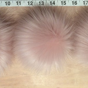 May include: Three fluffy, light pink faux fur pom-poms. The pom-poms are round and have a dense, textured appearance. They are arranged in a row on a wooden surface, with a measuring tape visible in the background.
