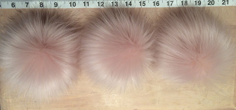 May include: Three fluffy, light pink faux fur pom-poms. The pom-poms are round and have a dense, textured appearance. They are arranged in a row on a wooden surface, with a measuring tape visible in the background.