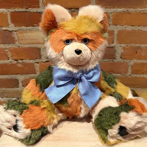 May include: A multi-coloured plush bear with a light blue bow. The bear's fur is a mix of orange, green, yellow, and cream. It has black paw pads and a black nose. The bear is positioned in front of a brick wall.