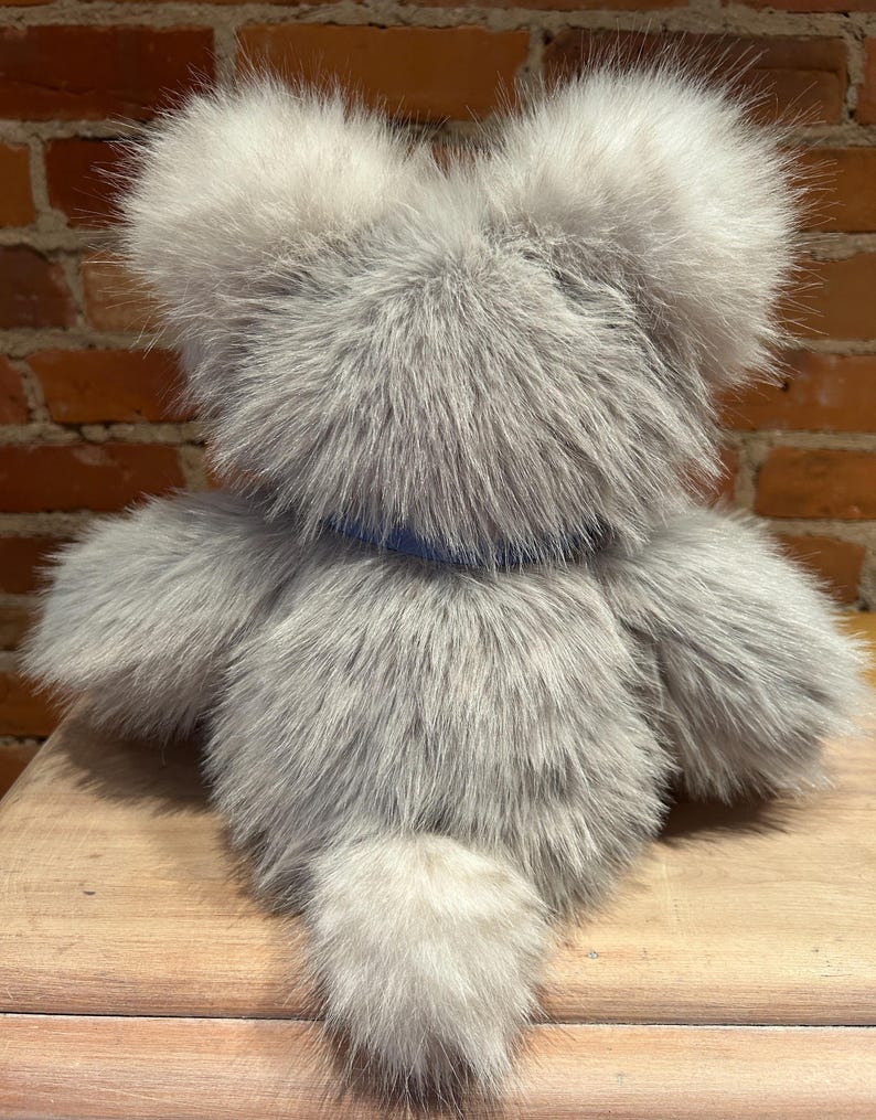 K&ouml;nnte beinhalten: Ein grauer Pl&uuml;schteddyb&auml;r mit einem langen, flauschigen Schwanz. Der B&auml;r tr&auml;gt ein blaues Halsband und sitzt auf einer Holzoberfl&auml;che.