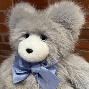 K&ouml;nnte beinhalten: Ein grauer Pl&uuml;schteddyb&auml;r mit wei&szlig;em Gesicht und blauer Schleife. Der B&auml;r hat schwarze Augen und eine schwarze Nase.