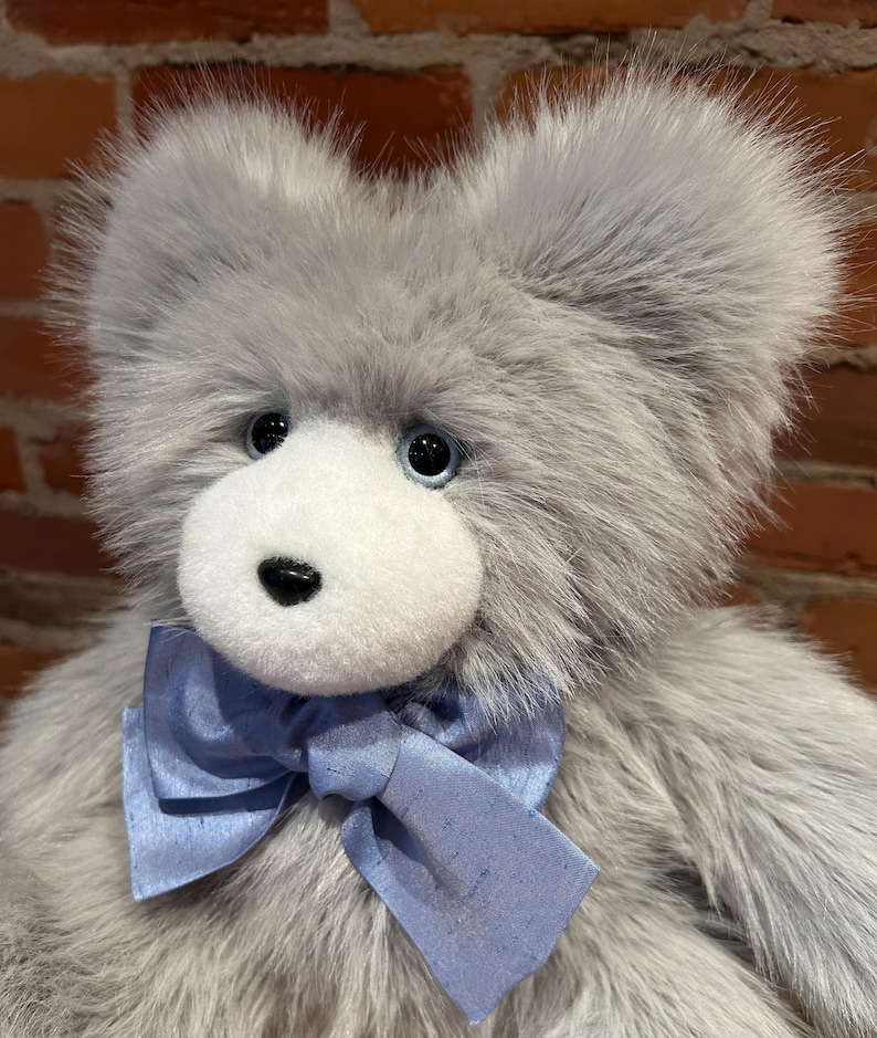 K&ouml;nnte beinhalten: Ein grauer Pl&uuml;schteddyb&auml;r mit wei&szlig;em Gesicht und blauer Schleife. Der B&auml;r hat schwarze Augen und eine schwarze Nase.