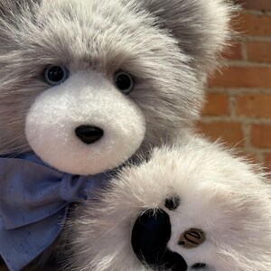 K&ouml;nnte beinhalten: Ein grauer und wei&szlig;er Pl&uuml;schteddyb&auml;r mit einer blauen Schleife. Der B&auml;r hat schwarze Augen und eine schwarze Nase. Die Pfote des B&auml;ren ist mit schwarzen Pfotenballen sichtbar.