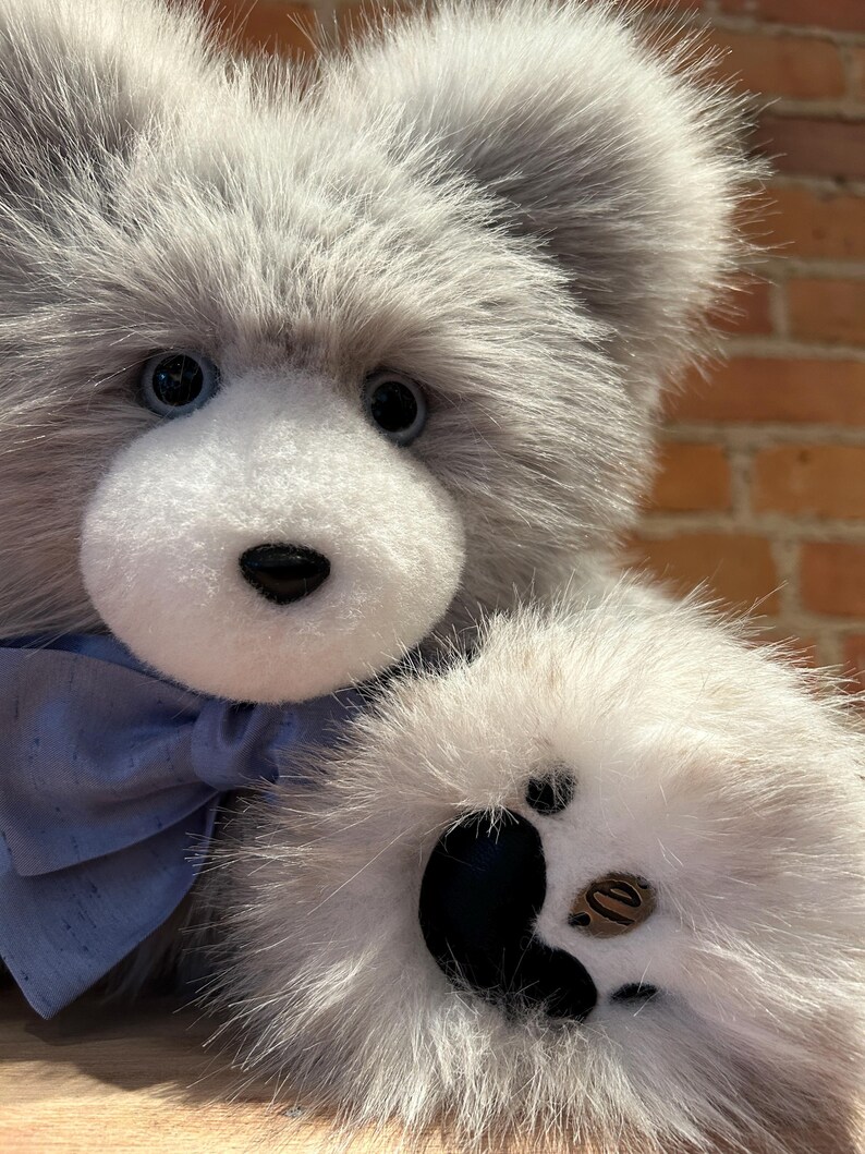 K&ouml;nnte beinhalten: Ein grauer und wei&szlig;er Pl&uuml;schteddyb&auml;r mit einer blauen Schleife. Der B&auml;r hat schwarze Augen und eine schwarze Nase. Die Pfote des B&auml;ren ist mit schwarzen Pfotenballen sichtbar.