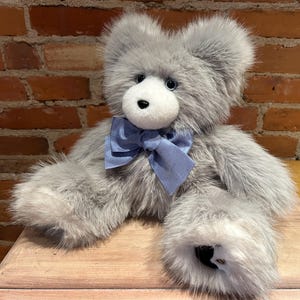 K&ouml;nnte beinhalten: Ein grauer Pl&uuml;schteddyb&auml;r mit einer blauen Schleife aus Satin. Der B&auml;r hat schwarze Augen und eine schwarze Nase.