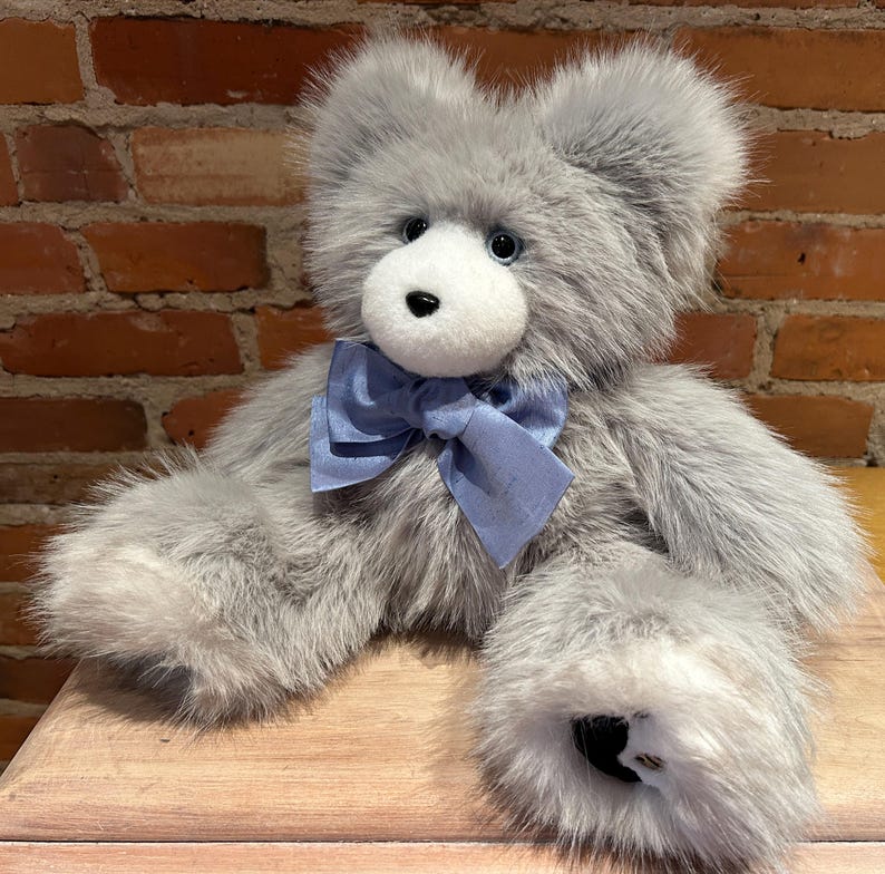 K&ouml;nnte beinhalten: Ein grauer Pl&uuml;schteddyb&auml;r mit einer blauen Schleife aus Satin. Der B&auml;r hat schwarze Augen und eine schwarze Nase.