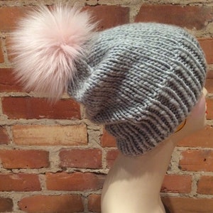 May include: A gray knit hat with a large pink faux fur pom-pom. The hat is made of thick yarn and has a ribbed brim. The hat is displayed on a mannequin head against a brick wall.
