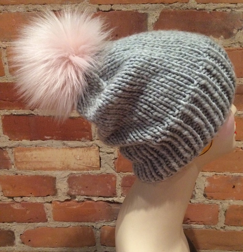 May include: A gray knit hat with a large pink faux fur pom-pom. The hat is made of thick yarn and has a ribbed brim. The hat is displayed on a mannequin head against a brick wall.