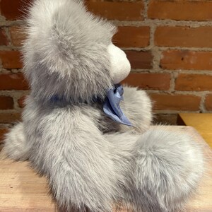 K&ouml;nnte beinhalten: Ein grauer Pl&uuml;schteddyb&auml;r mit einem blauen Band um den Hals. Der B&auml;r sitzt auf einer Holzoberfl&auml;che.