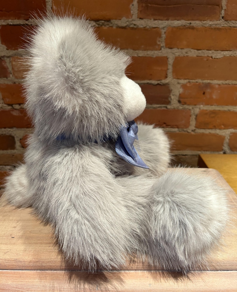 K&ouml;nnte beinhalten: Ein grauer Pl&uuml;schteddyb&auml;r mit einem blauen Band um den Hals. Der B&auml;r sitzt auf einer Holzoberfl&auml;che.