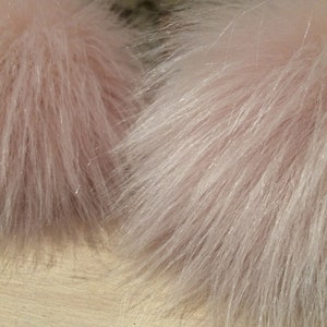 May include: Close-up of two fluffy, light pink faux fur pom-poms. The fur has a soft, textured appearance with long, delicate strands. The pom-poms are likely for crafting or fashion accessories.