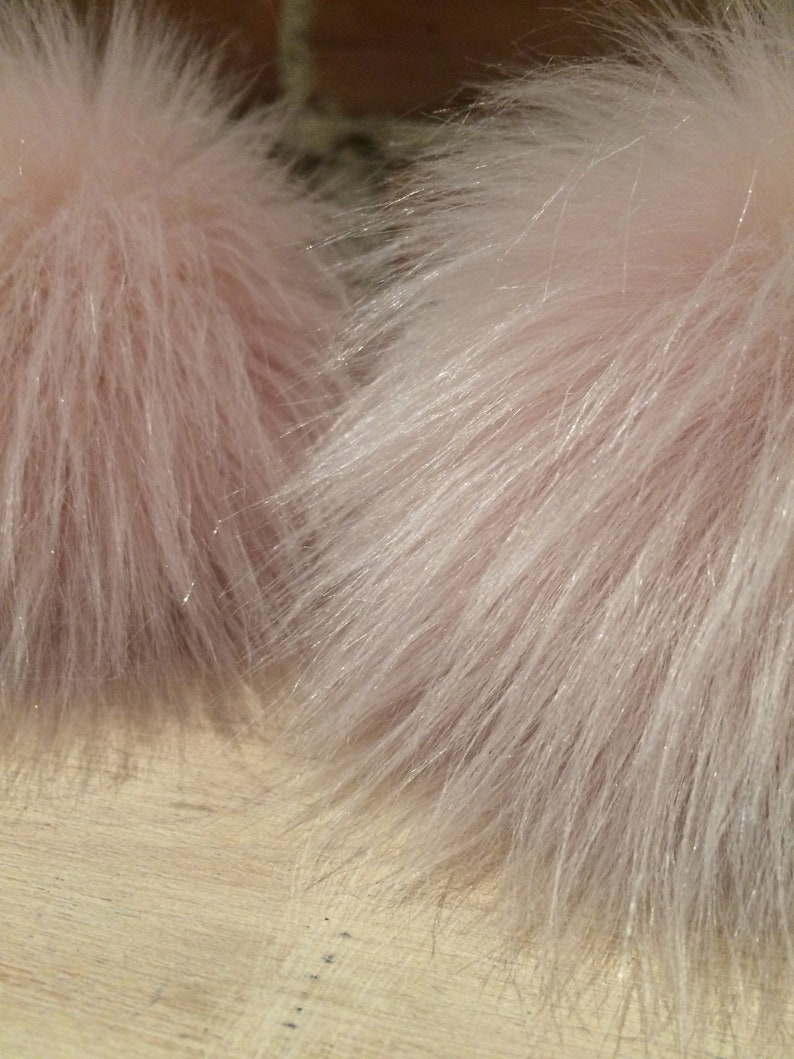 May include: Close-up of two fluffy, light pink faux fur pom-poms. The fur has a soft, textured appearance with long, delicate strands. The pom-poms are likely for crafting or fashion accessories.